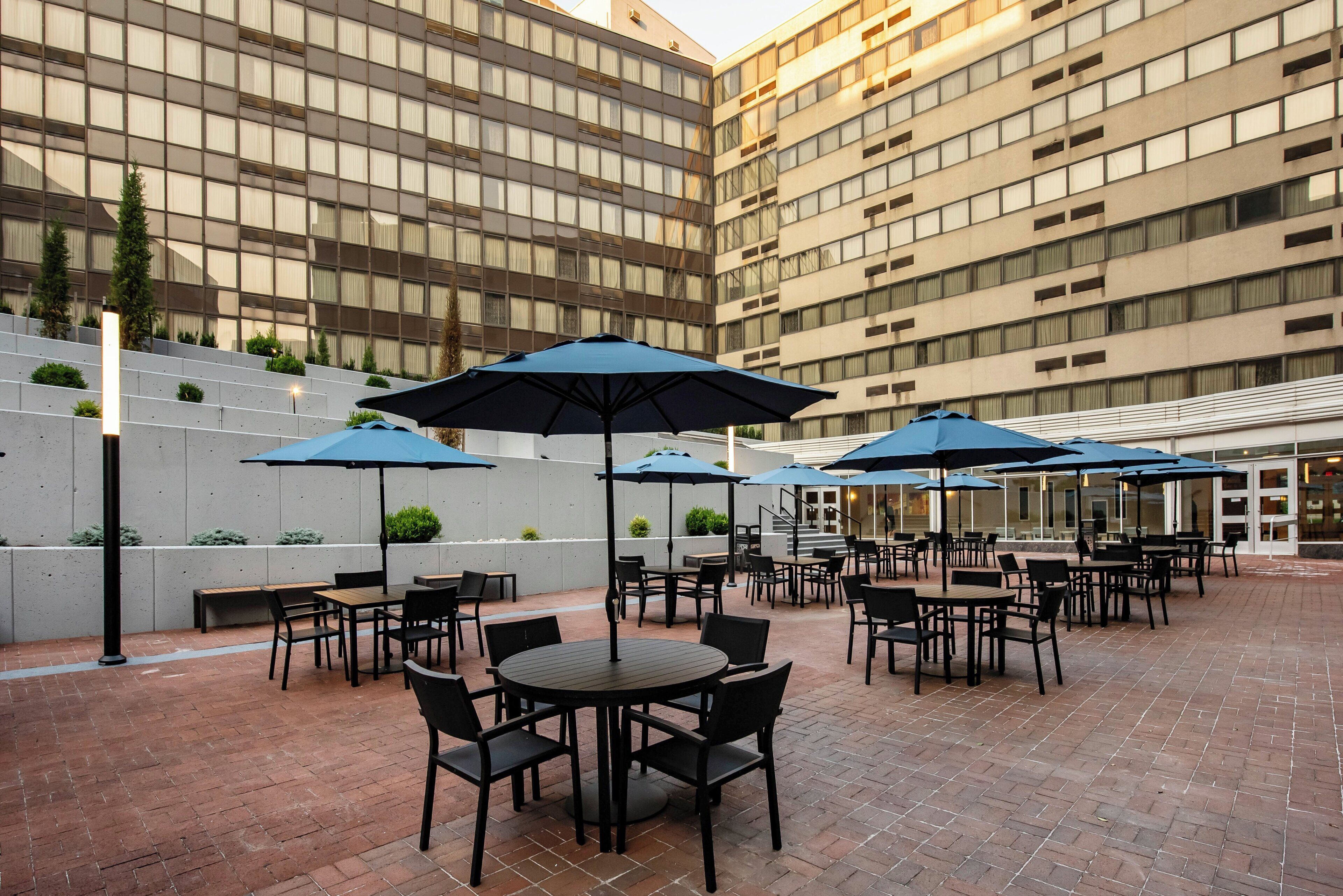 Ein Außenbereich mit Terrasse, ausgestattet mit runden Tischen und Stühlen, blauen Sonnenschirmen und einem Pflasterboden.