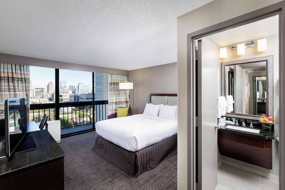 Habitación de hotel con una cama grande, un escritorio con computadora, un baño con espejo y lavamanos, y vista a la ciudad.