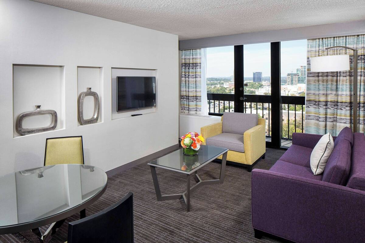 Una habitación de hotel moderna con una mesa de centro de vidrio, un sofá púrpura, un sillón amarillo, un televisor montado en la pared y un ventanal con vistas a la ciudad.