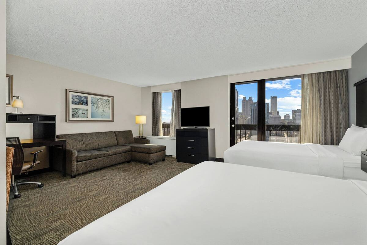 Habitación de hotel con cama, sofá, escritorio con ordenador, televisión y vistas a la ciudad.