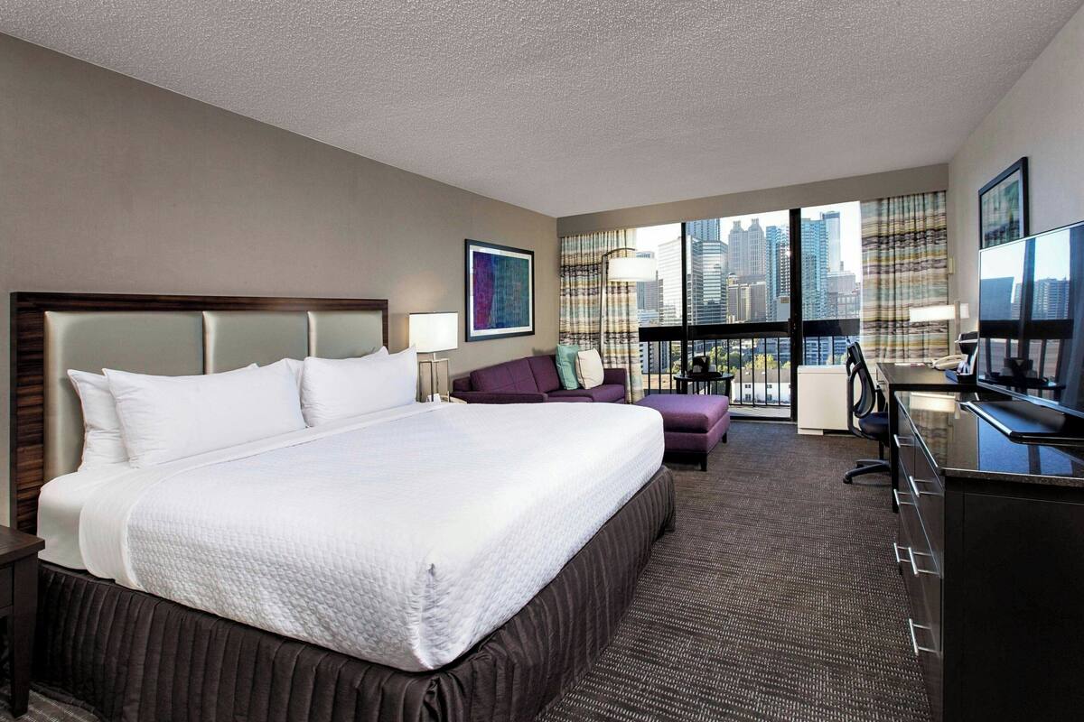 Habitación de hotel con una cama grande, un escritorio, una silla, un sofá y vistas de la ciudad a través de amplias ventanas.