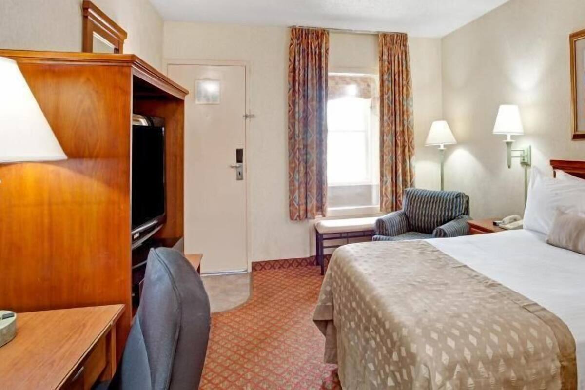 Quarto de hotel com cama, escrivaninha, cadeira, televisão e um quadro na parede.
