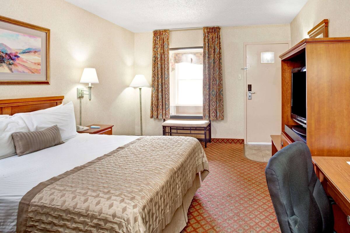 Quarto de hotel com uma cama grande, uma secretária com cadeira, televisão e janela com cortinas.