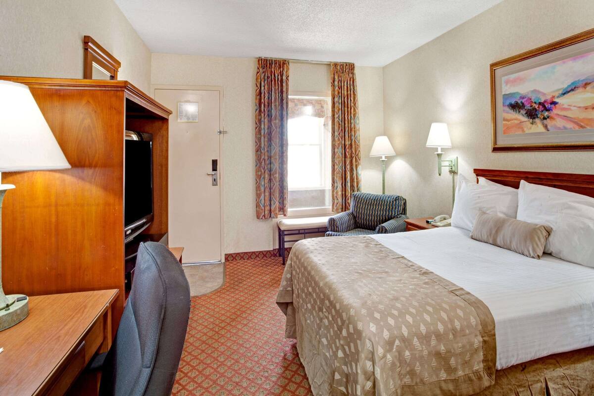 Quarto de hotel com cama, escrivaninha, cadeira, televisão e janela com cortinas.