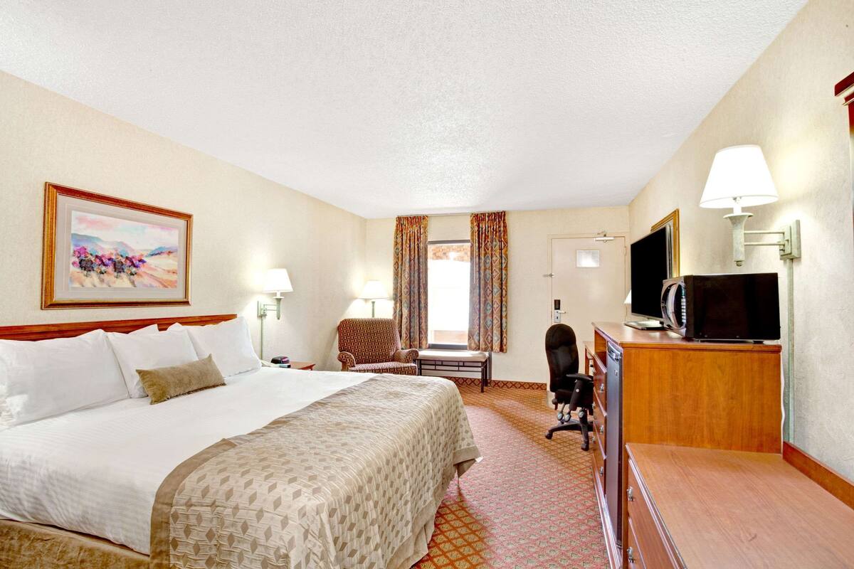 Quarto de hotel com uma cama grande, uma escrivaninha com televisão, uma cadeira e um abajur.