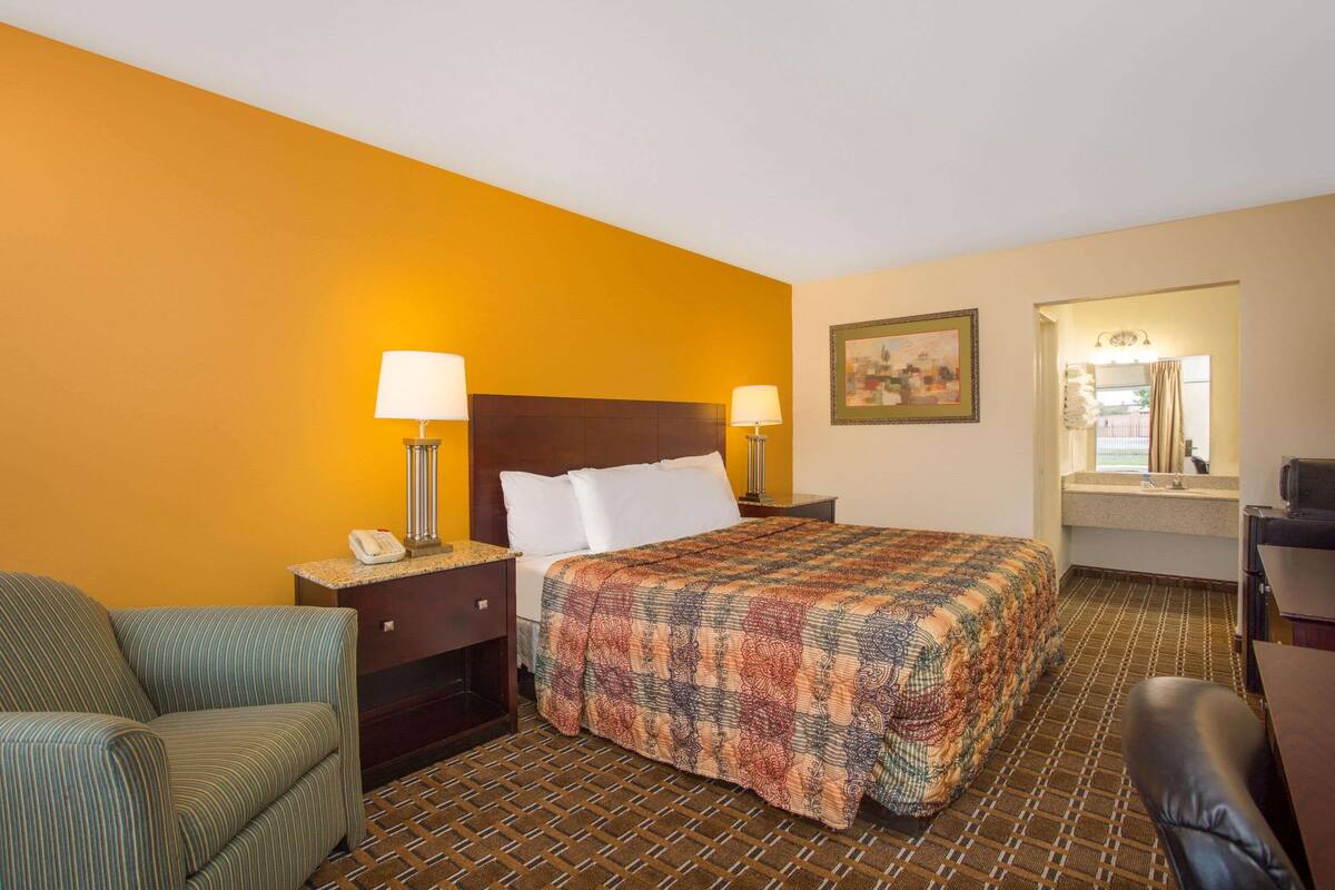 A hotel room with a bed, two bedside lamps, a chair, a desk, and a bathroom visible through an open door.