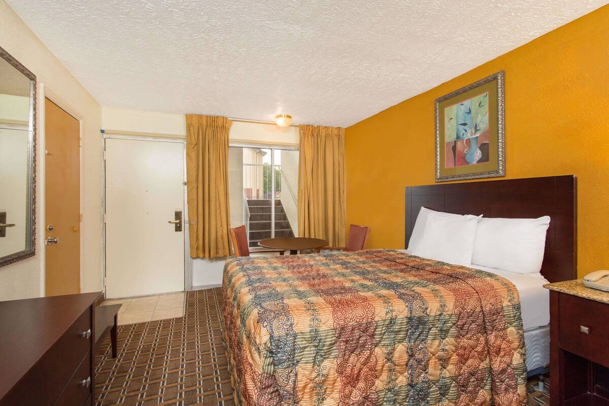 A hotel room with a bed, a desk, a chair, a painting on the wall, and a view of the outside.
