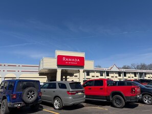 Un parking où sont garés plusieurs VUS, un bâtiment sur lequel on peut lire l’inscription « RAMADA BY WYNDHAM », et un ciel bleu dégagé.