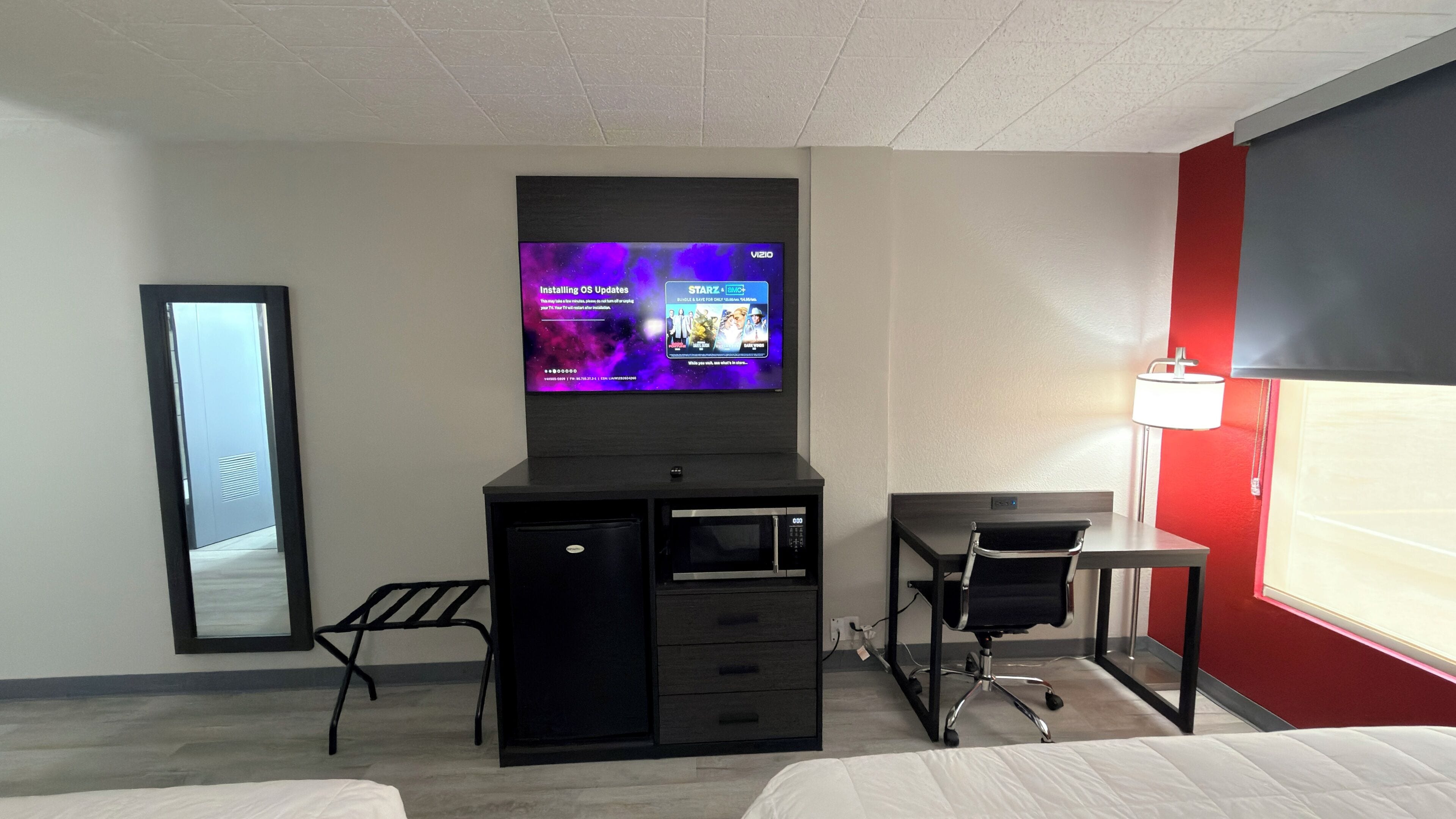 A hotel room with a TV mounted on the wall, a desk with a chair, a bed, and a small table with a lamp.