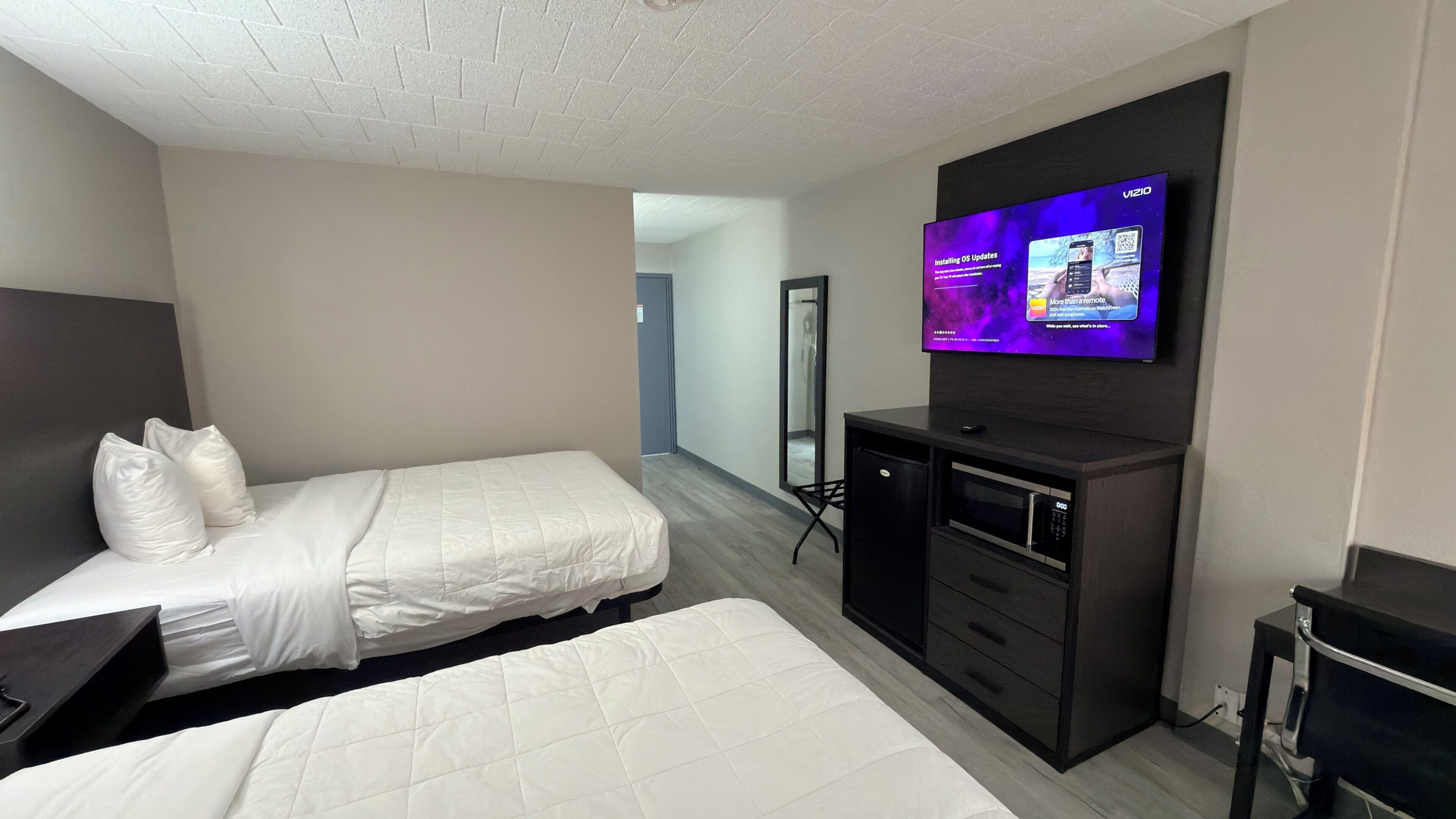 Habitación de hotel con dos camas, televisor montado en la pared, un espejo, escritorio y una kitchenette pequeña.