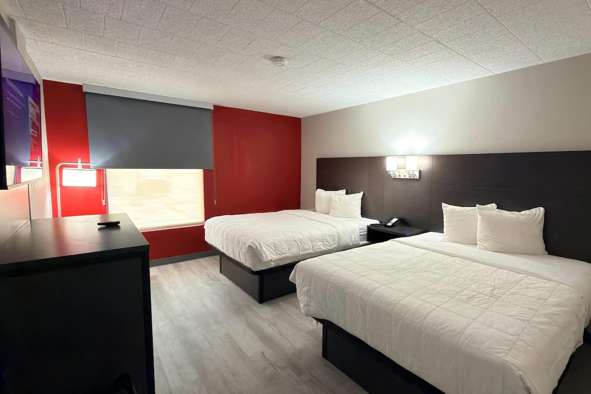 A hotel room with two beds, a desk, a red accent wall, and a window.