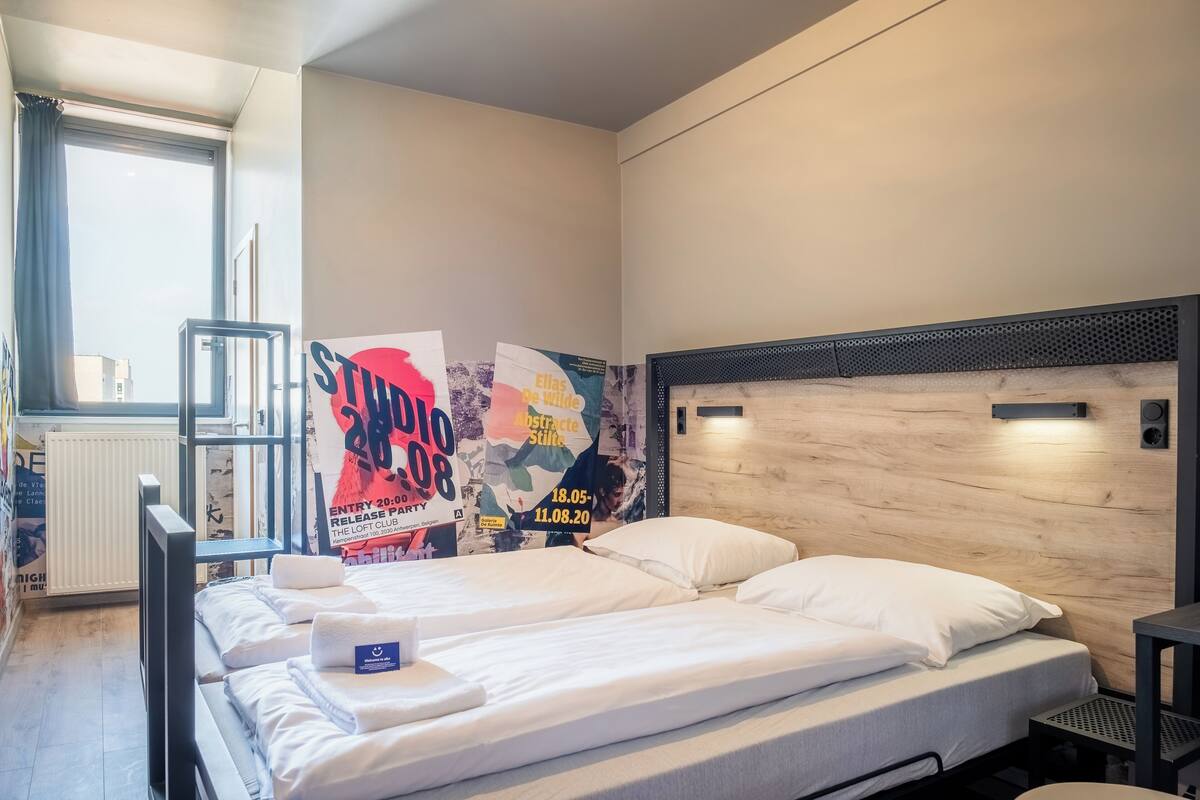 A hotel room with a bed, a small table, and a radiator. There are posters on the wall advertising an event.
