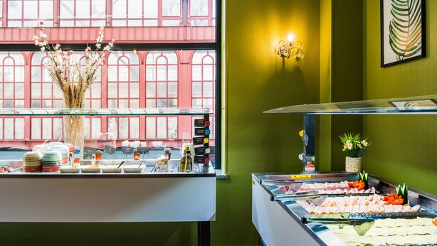 A buffet area with a window view of a red building, a vase with dried flowers, and a wall-mounted light fixture. - a&o Antwerpen Centraal (Antwerp)