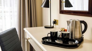 Une chambre d’hôtel avec un bureau, deux tasses noires, un plateau avec une bouilloire et une cuillère, une lampe et une fenêtre avec des rideaux.