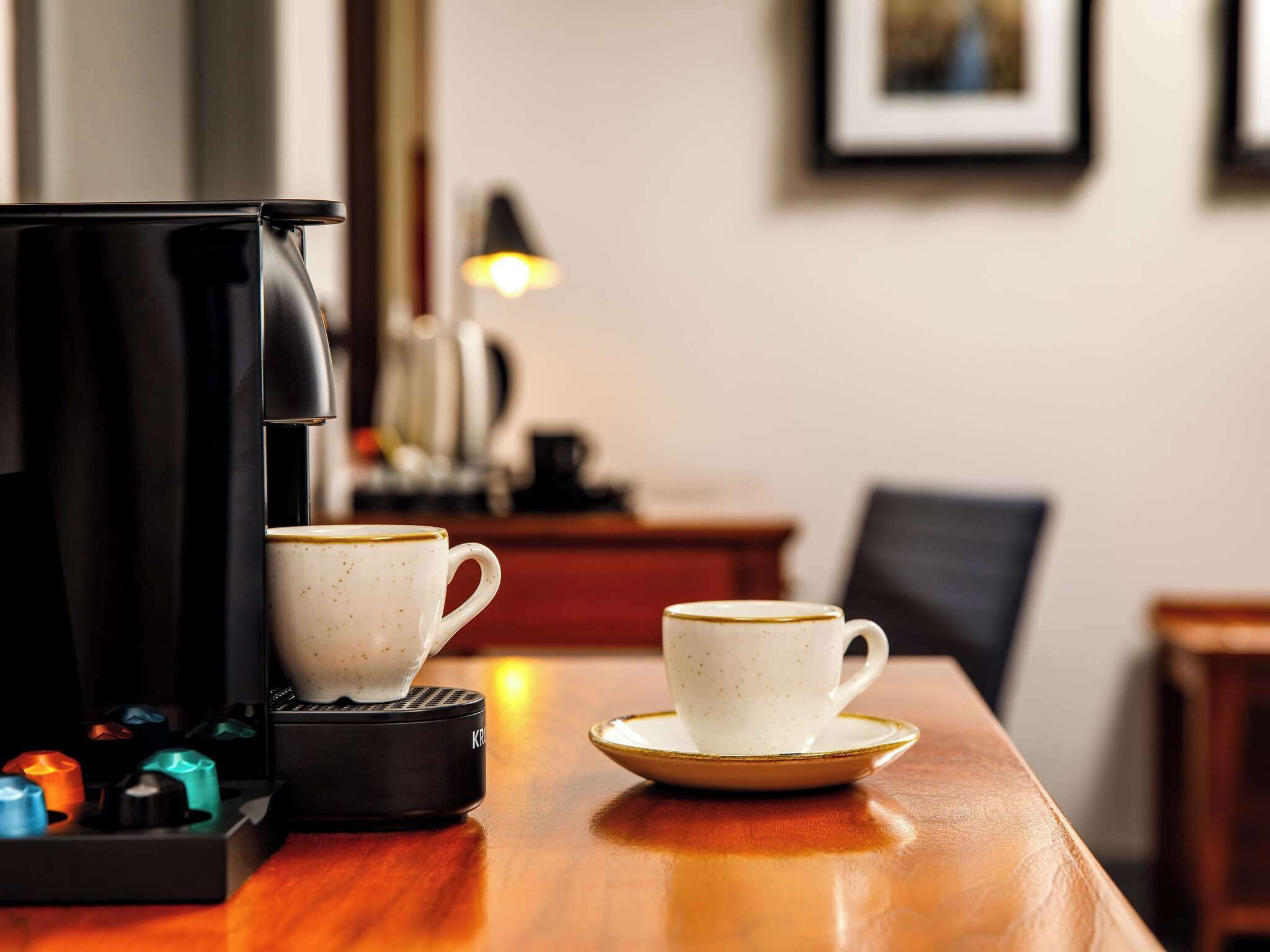 A coffee machine, two cups, and a small tray on a wooden table in a room with a lamp and framed picture on the wall.