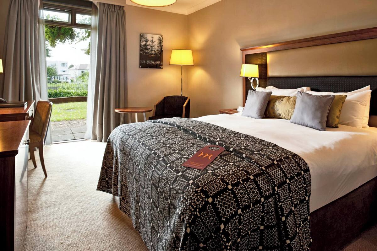 Ein Hotelzimmer mit einem großen Bett, zwei Nachttischlampen, einem Schreibtisch mit Stuhl, einem kleinen Tisch und Blick ins Freie.
