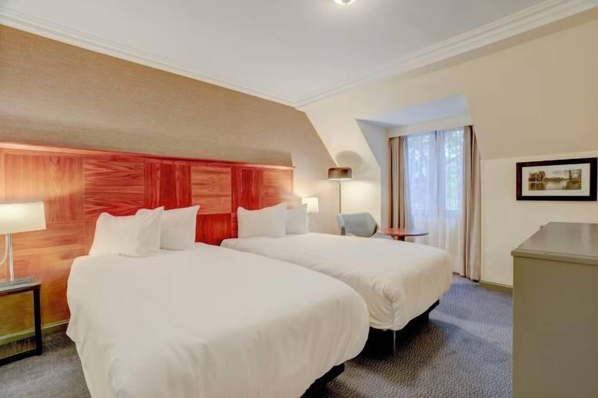 Ein Hotelzimmer mit zwei Betten, einem Schreibtisch, einer Lampe und einem Fenster mit Vorhängen.