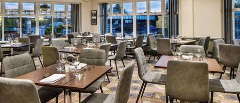 Een restaurant in een hotel, met meerdere tafels en stoelen, grote ramen en een kamerbreed tapijt met een patroon.