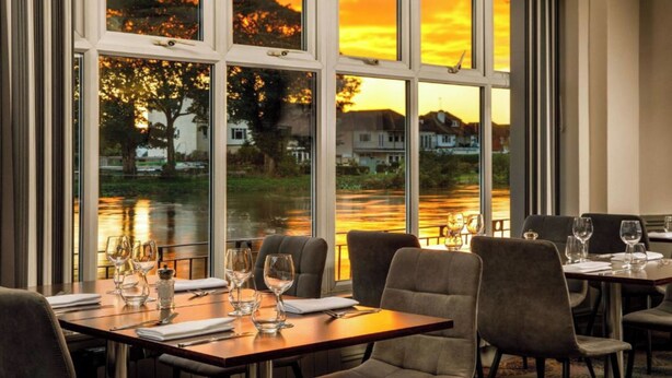 Un restaurant offrant une vue sur un coucher de soleil se reflétant sur une étendue d’eau, avec de grandes fenêtres et des tables dressées pour le dîner.
