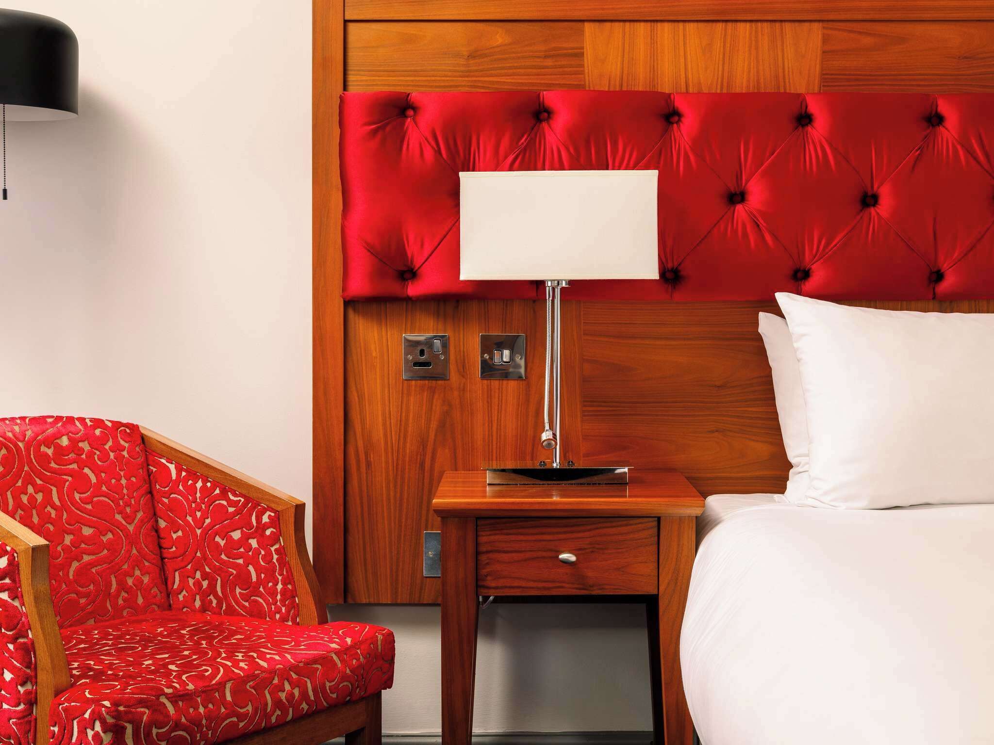 Habitación de hotel con un cabecero tapizado rojo, una mesita de noche con lámpara, una mesita auxiliar de madera, un sillón rojo estampado y una cama blanca.