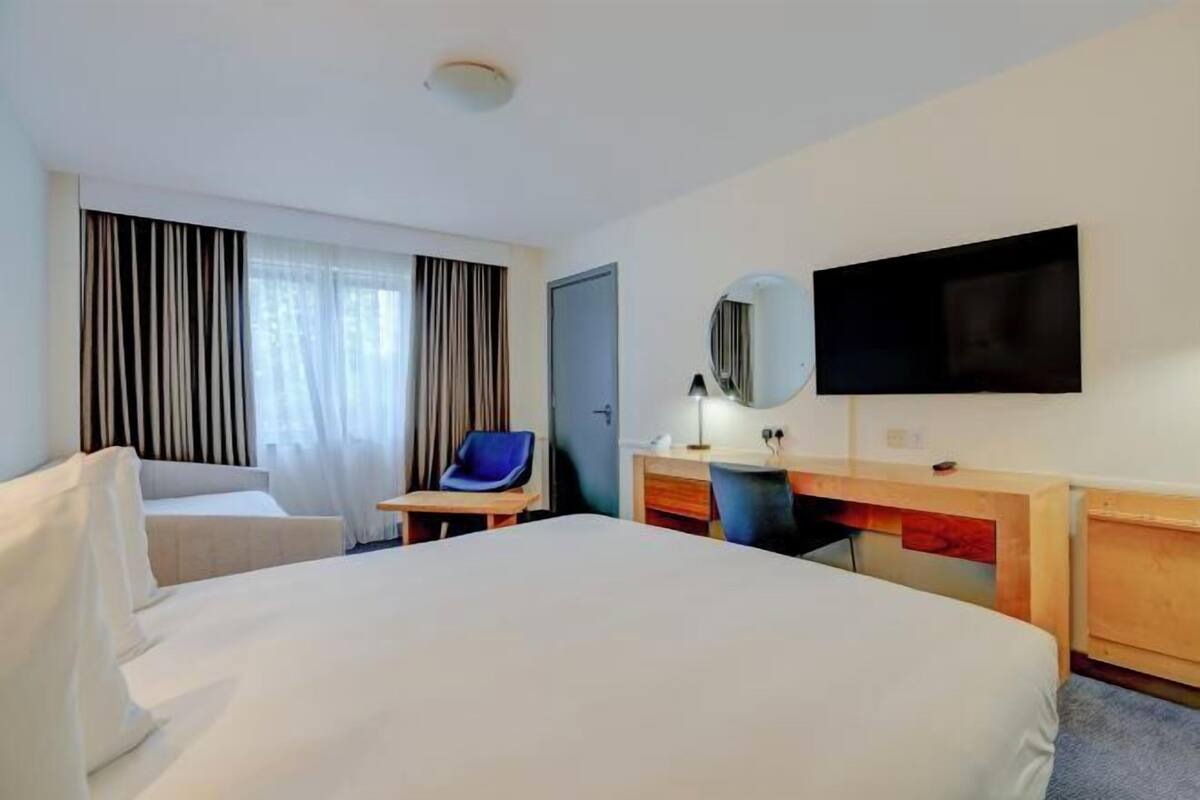 Ein Hotelzimmer mit einem großen Bett, einem Schreibtisch mit Stuhl, einem Fernseher und einem Fenster mit Vorhängen.