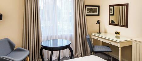 A hotel room with a bed, a desk, two chairs, a round table, a lamp, a mirror, and a window with curtains.