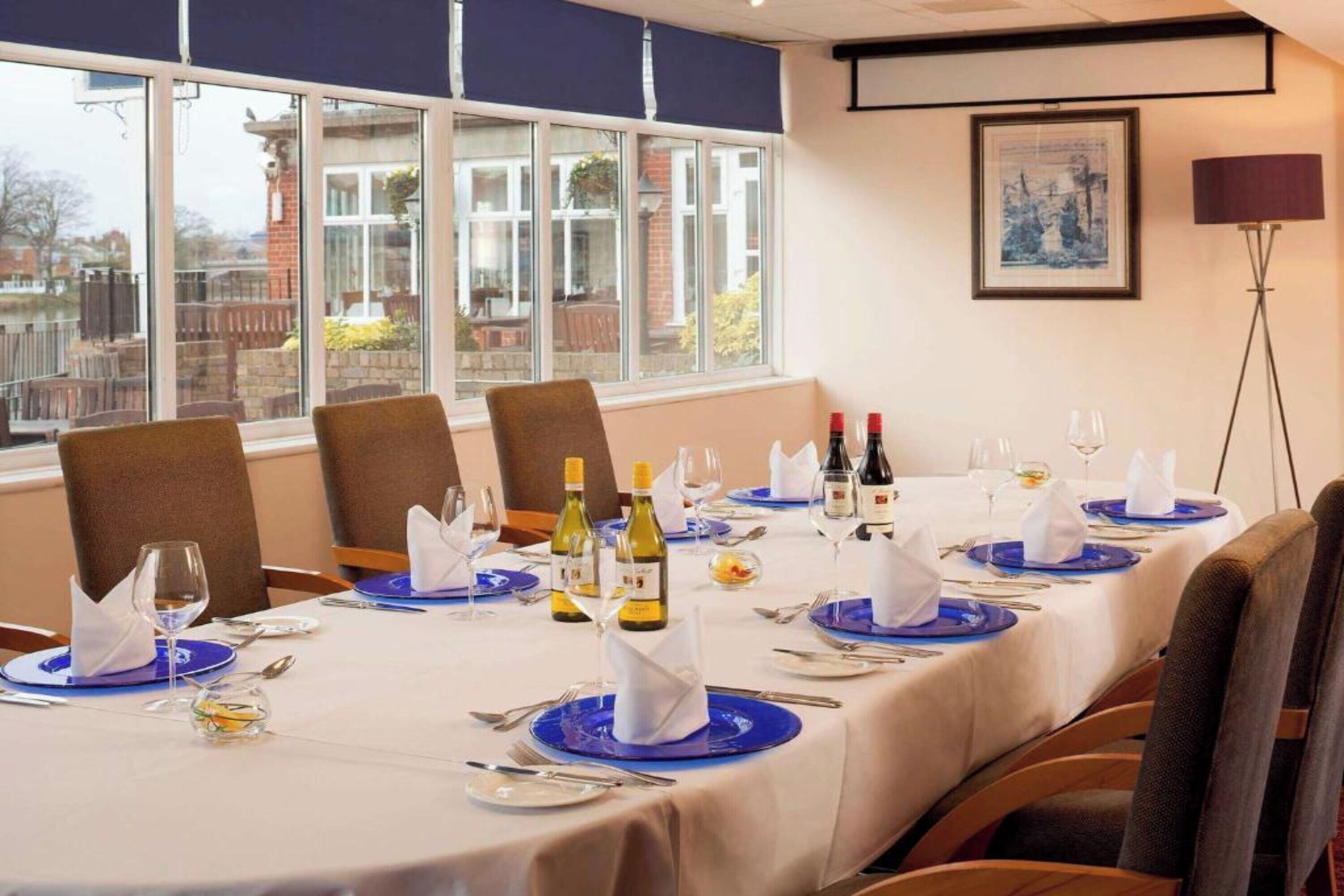 A dining area with a long table set for a meal, chairs, and a view of the outside through large windows.