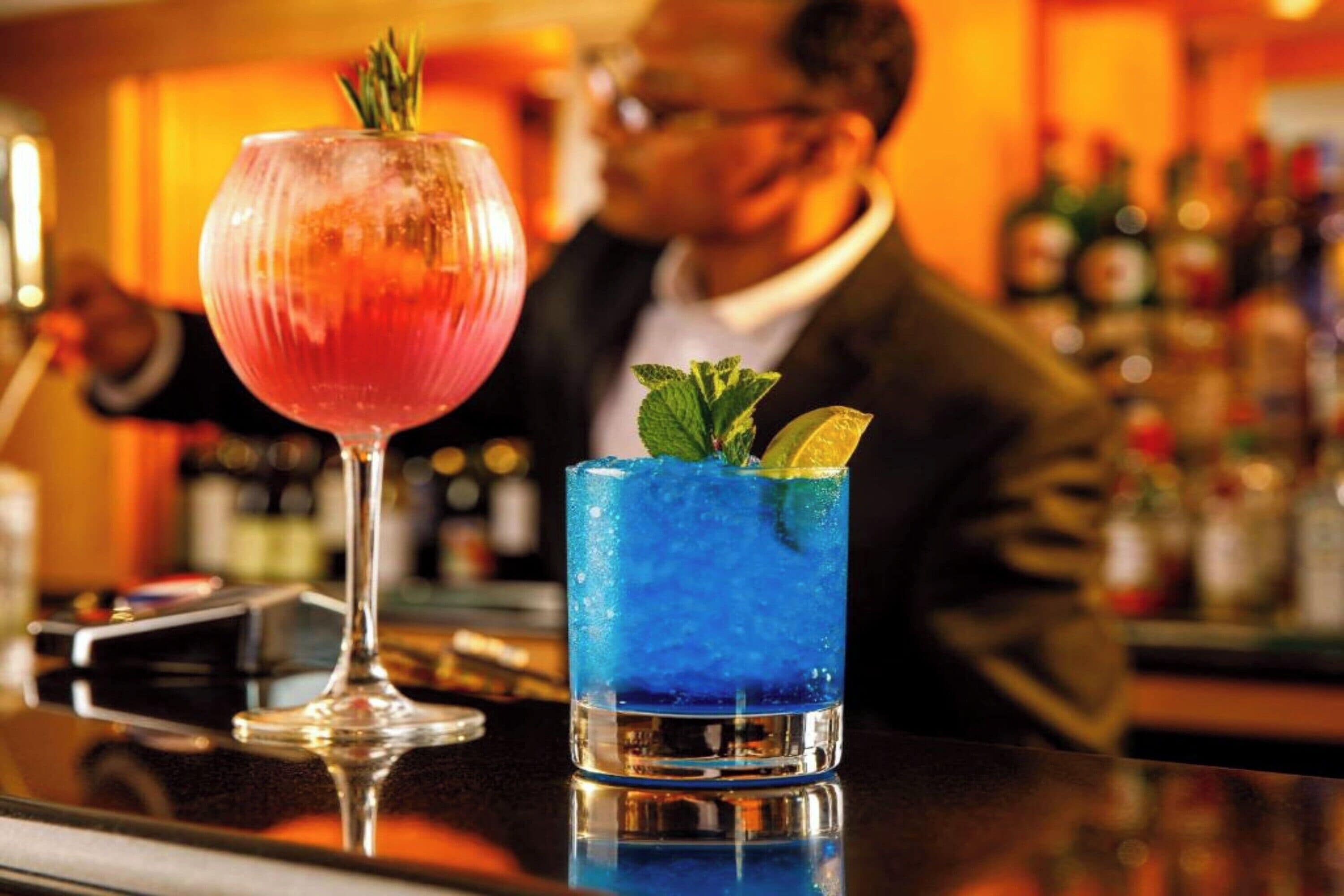Two cocktails on a bar counter with a blurred background of a bar and bottles.