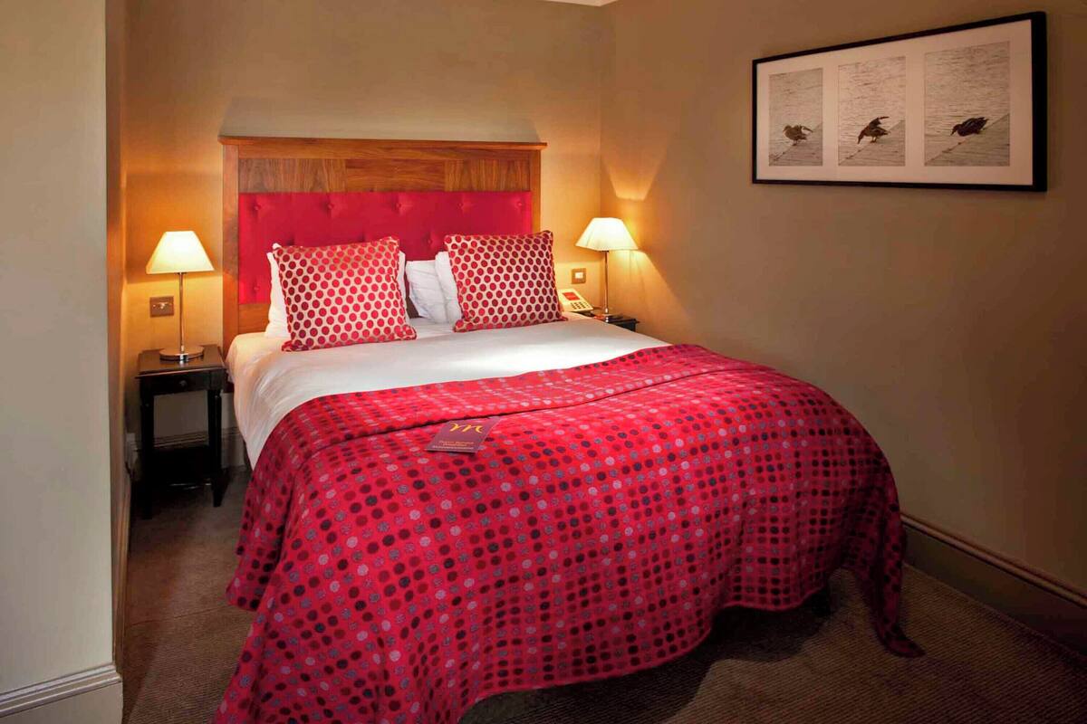 Ein Hotelzimmer mit einem großen Bett, rot-weißer Bettwäsche, zwei Nachttischlampen, einem gerahmten Bild an der Wand und einem kleinen Tisch mit einer Lampe.