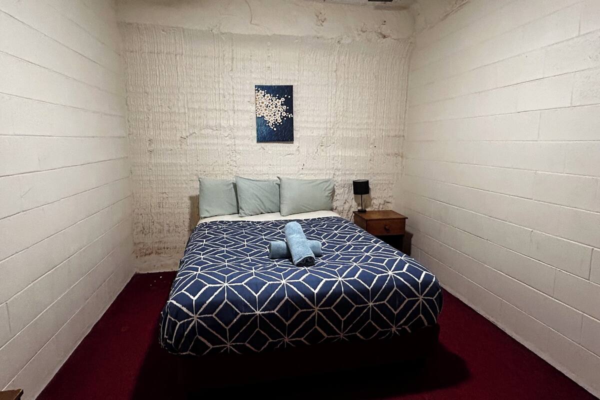 A narrow room with white brick walls, a bed with a geometric patterned bedspread, a small wooden nightstand with a lamp, and a wall-mounted artwork.