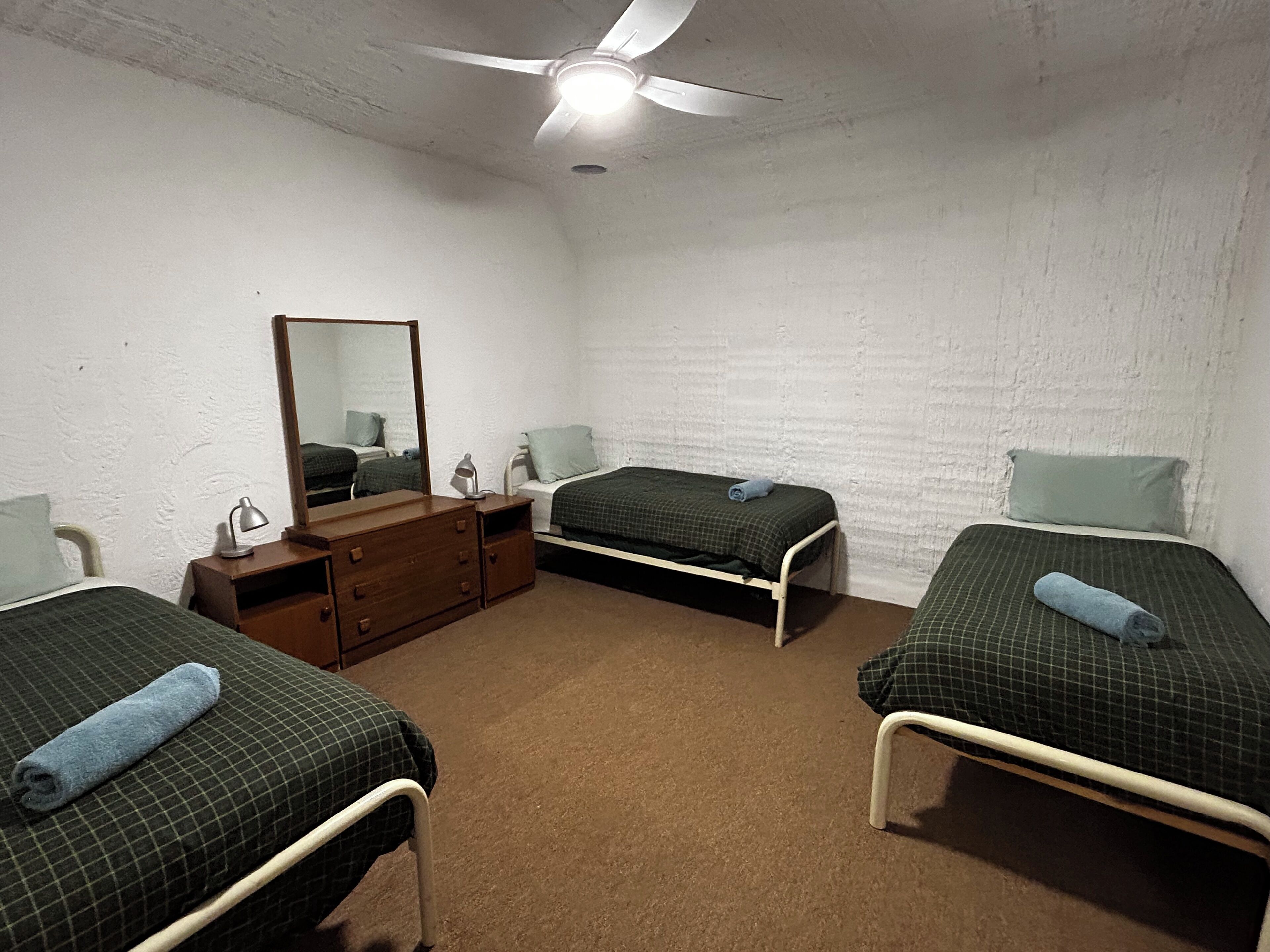 A small, simple room with three single beds, a wooden dresser, a mirror, and a ceiling fan.