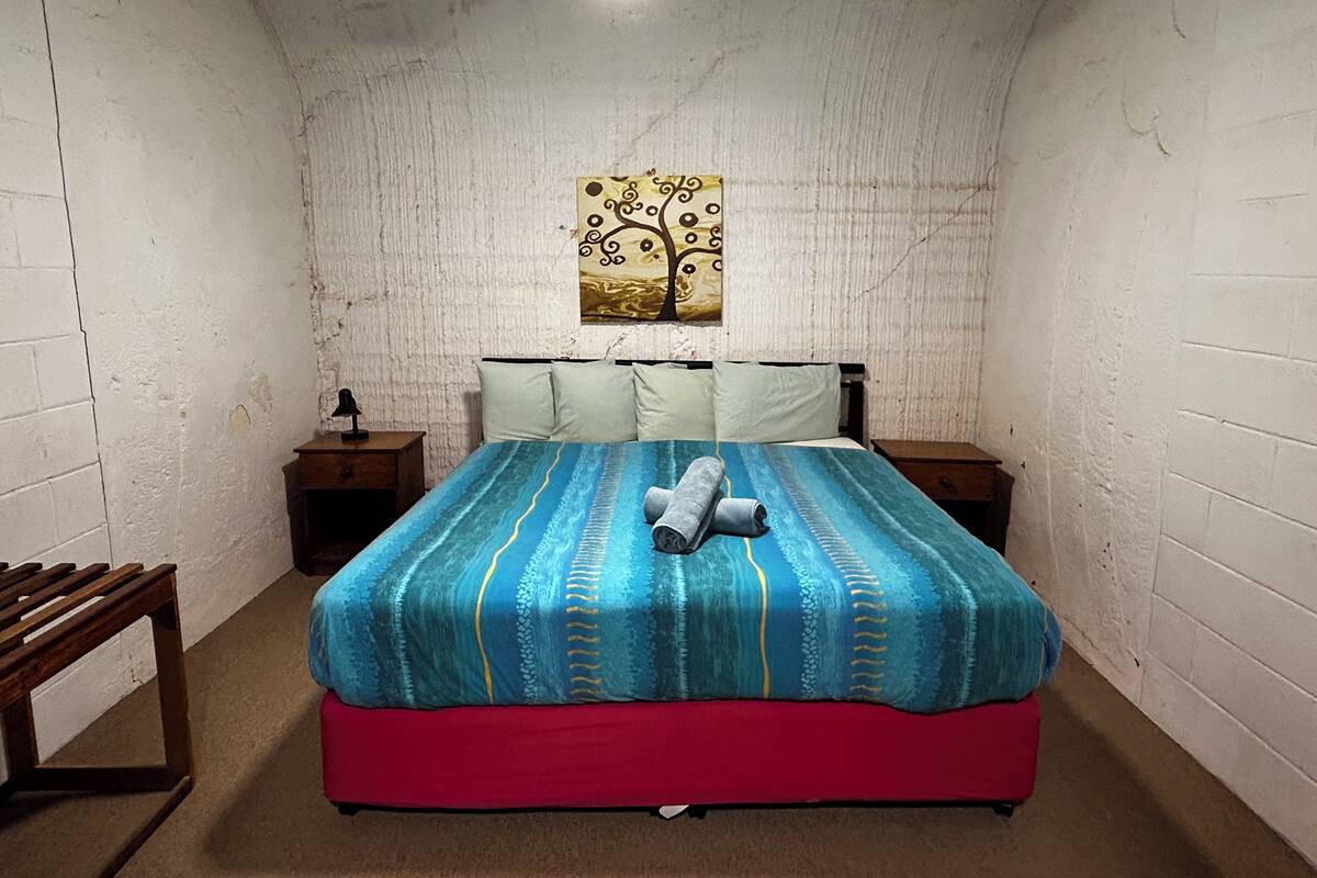 A small, narrow room with white brick walls, a single bed with a blue and red bedspread, a ceiling fan, and a small table with a lamp.