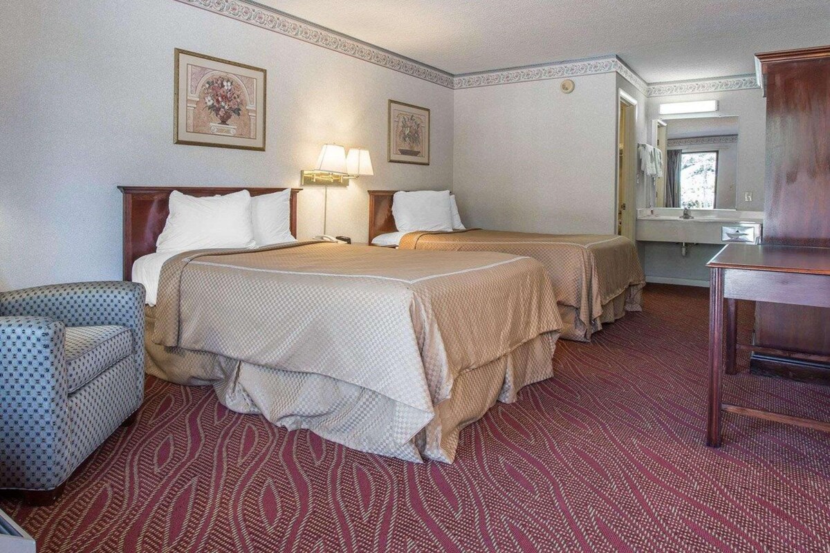 Quarto de hotel com duas camas, uma escrivaninha, uma cadeira, um abajur e um espelho.