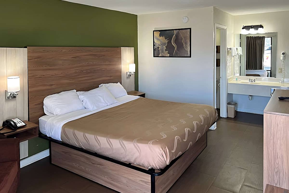 Une chambre d’hôtel comprenant un lit, une table de chevet, une lampe et une salle de bain avec un miroir et un lavabo.