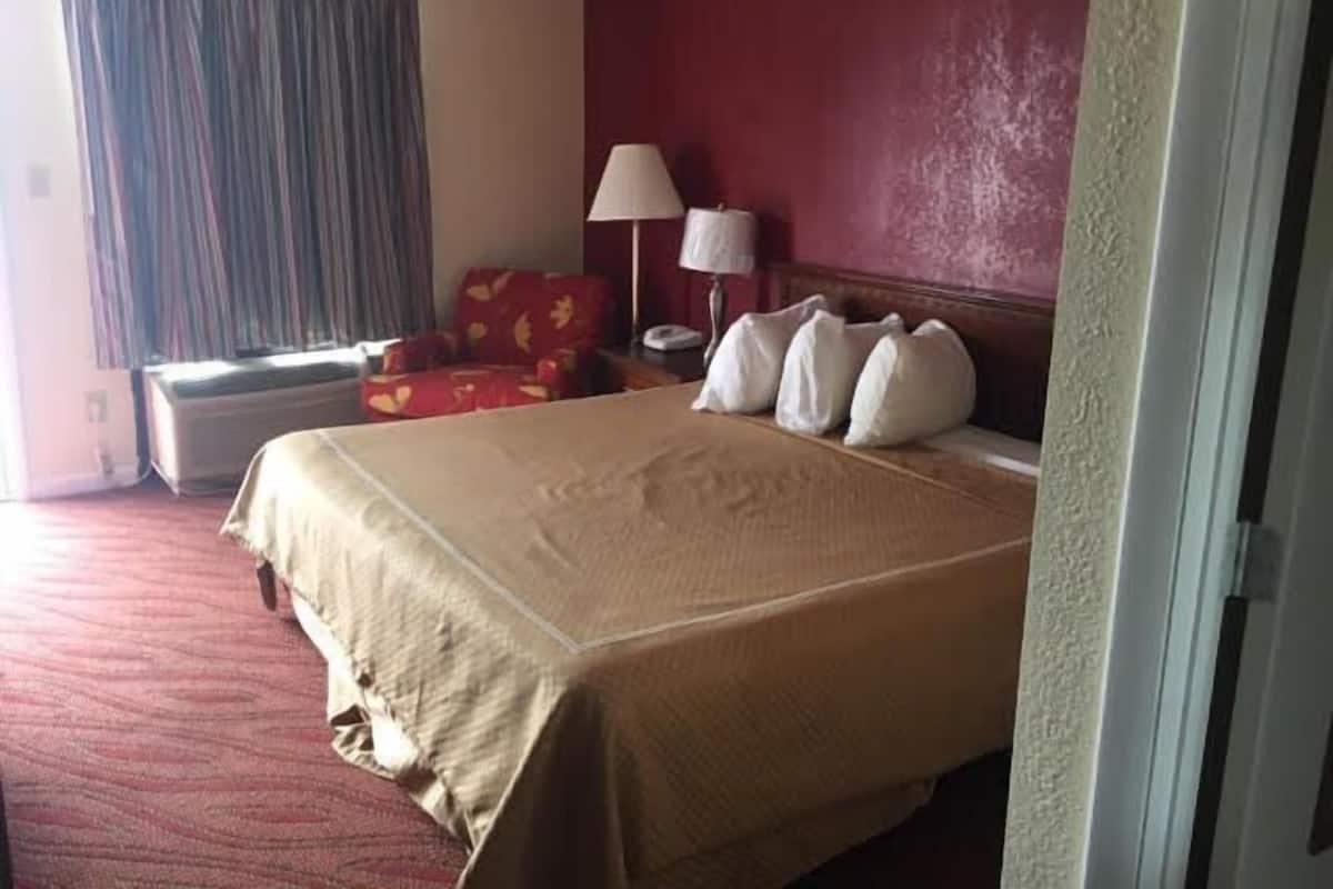 Une chambre d’hôtel avec un grand lit, un fauteuil rouge, une lampe et une fenêtre avec des rideaux.