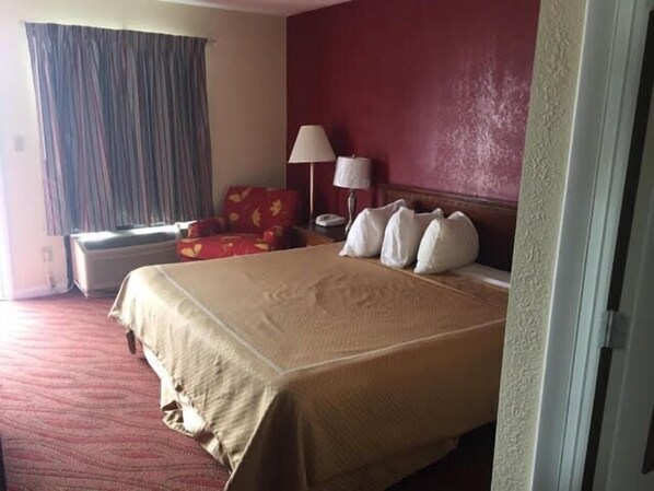 Habitación de hotel con una cama grande, un sillón rojo, una lámpara y un ventanal con cortinas.