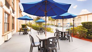 Un patio al aire libre con mesas y sillas bajo sombrillas azules, una piscina y un edificio con grandes ventanales.