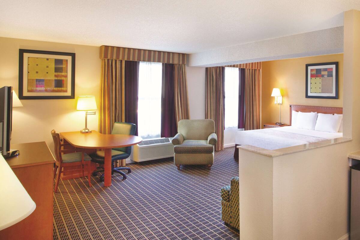 A hotel room with a bed, a desk with a chair, a small table, a lamp, an air conditioner, and curtains.