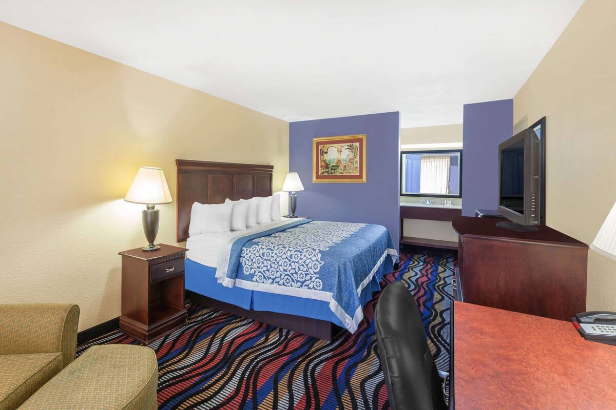 A hotel room with a bed, two bedside lamps, a desk with a computer, a television, and a patterned carpet.