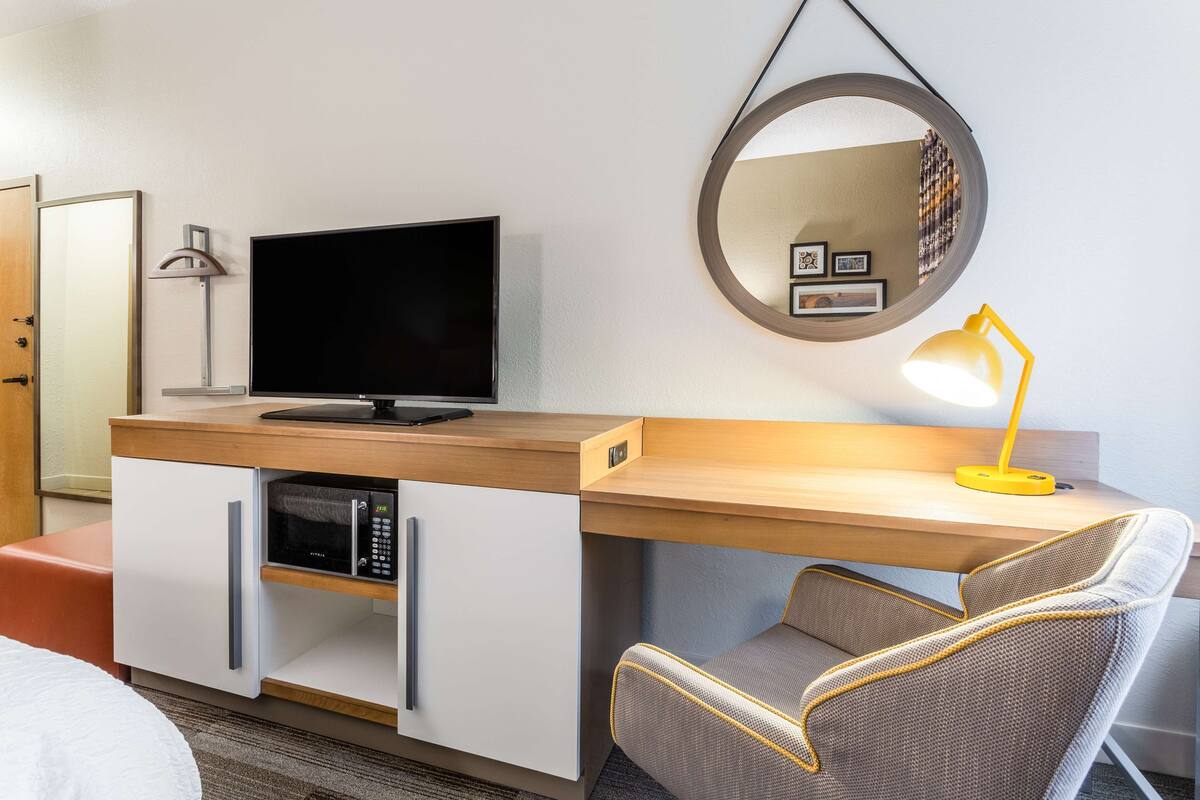 A hotel room with a wooden desk, a flat-screen TV, a microwave, a yellow desk lamp, a round mirror, and a comfortable chair.