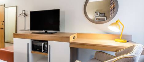 A hotel room with a wooden desk, a flat-screen TV, a microwave, a yellow desk lamp, a round mirror, and a comfortable chair.