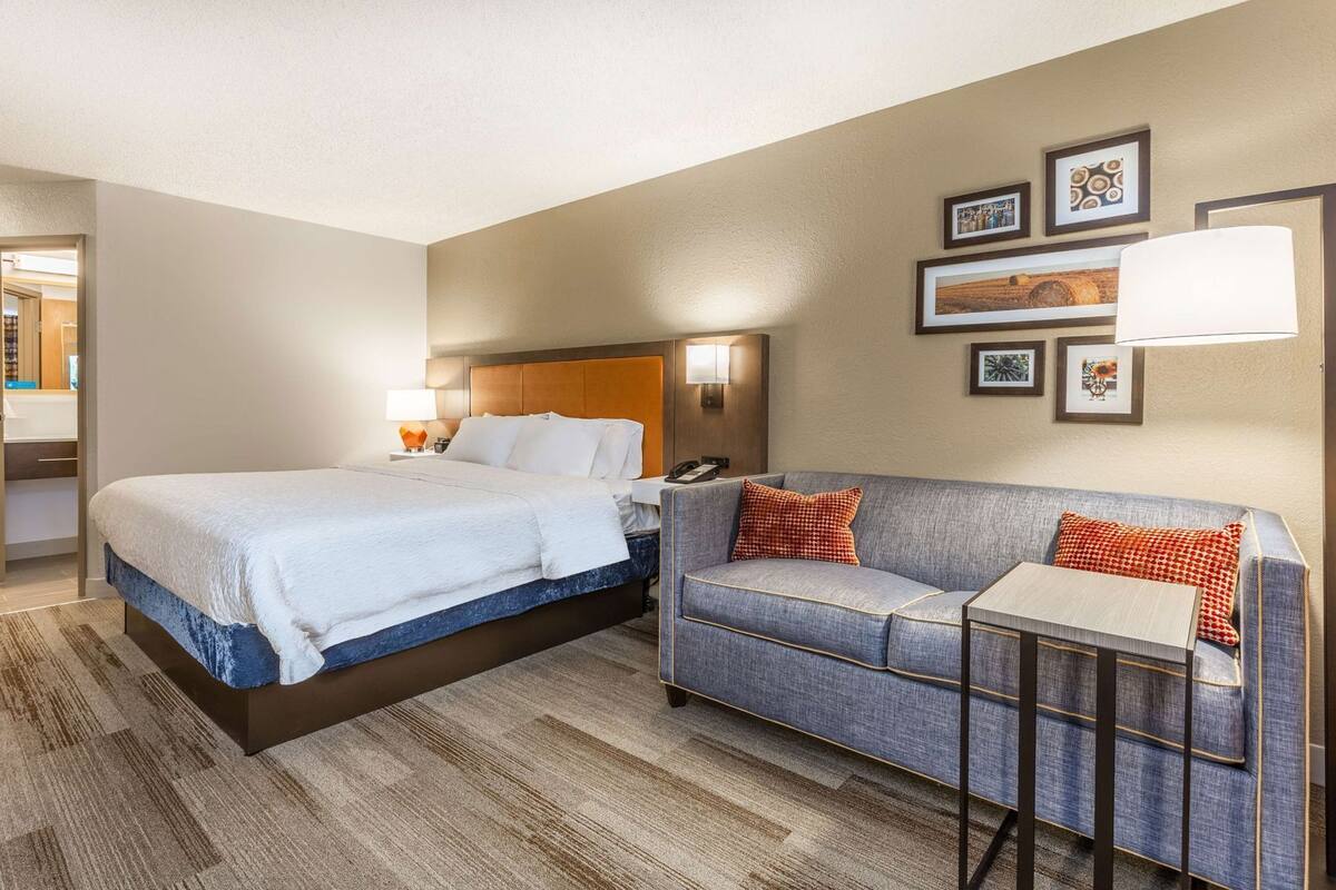 A hotel room with a bed, a sofa, a small table, and wall-mounted artwork.