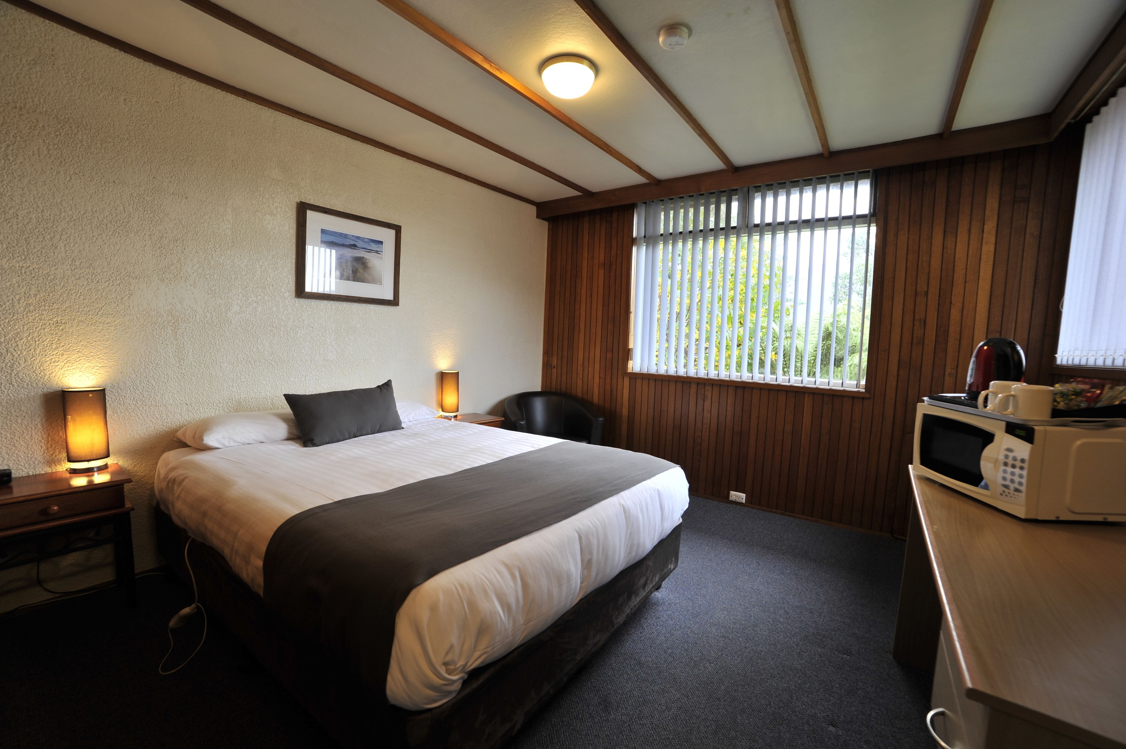 A hotel room with a large bed, bedside lamps, a wooden ceiling, a window with blinds, and a microwave on a wooden table.