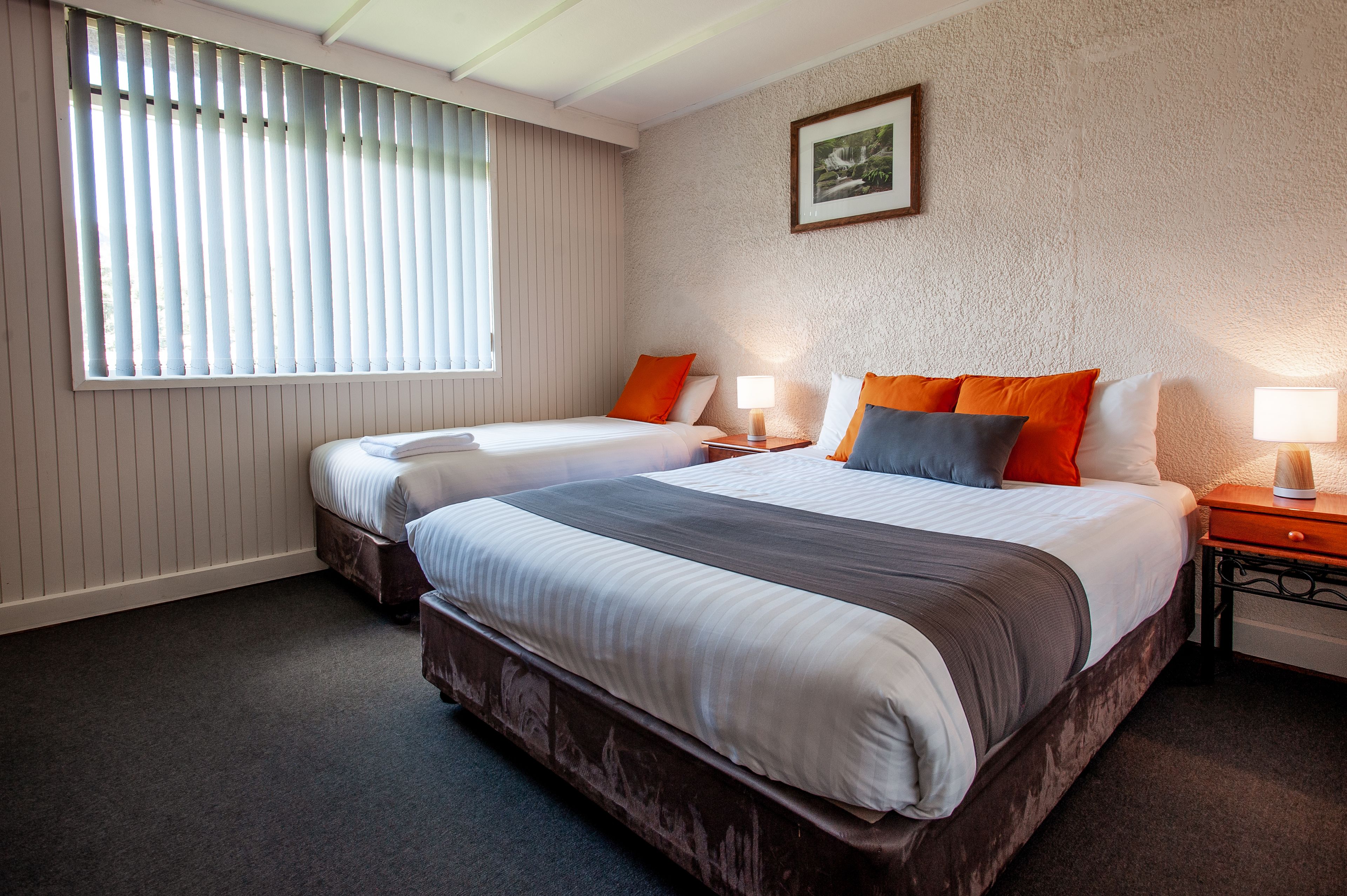 A hotel room with two beds, a large window with blinds, a framed picture on the wall, and a nightstand with a lamp.