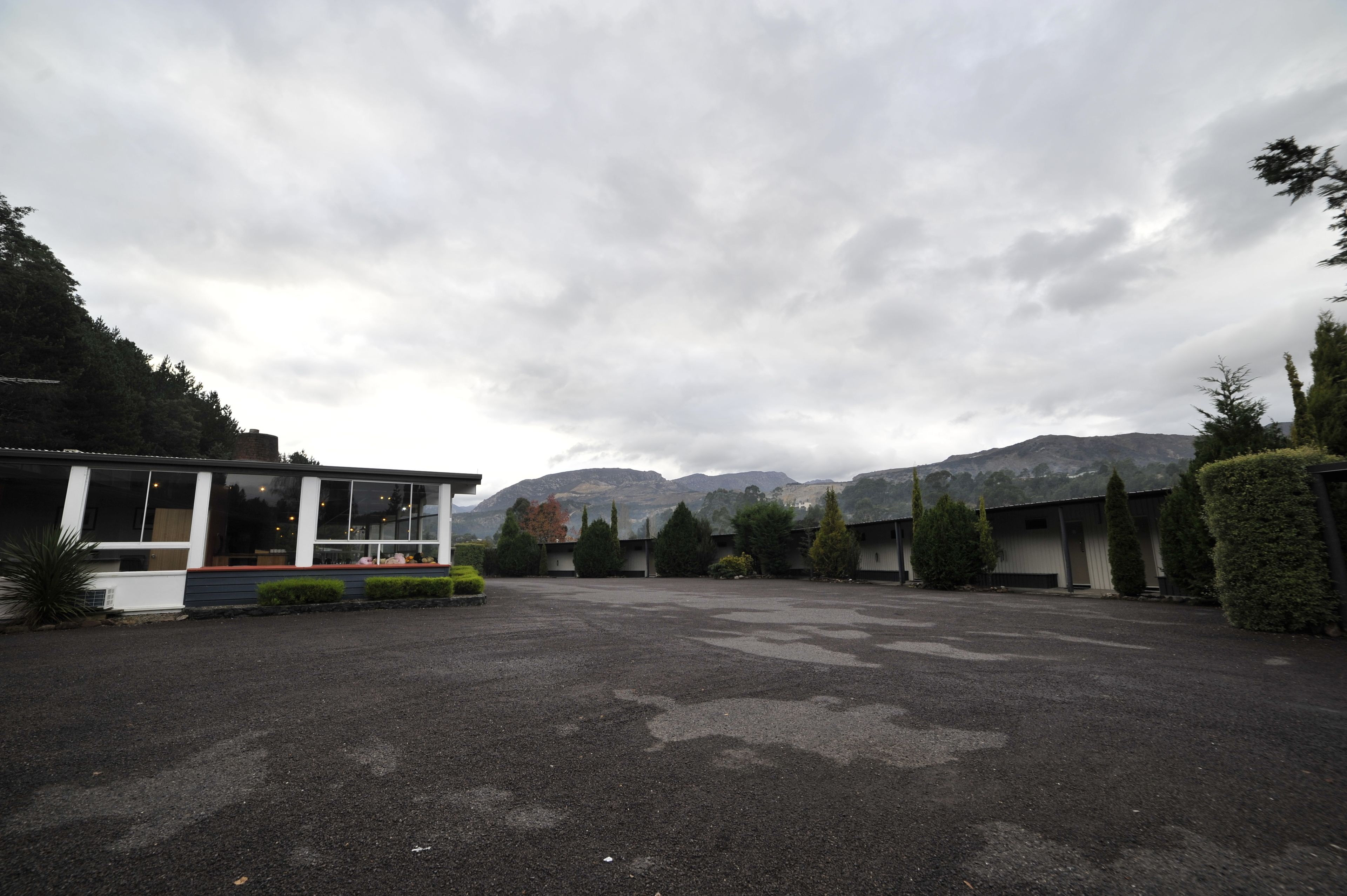 En parkeringsplats med en modern byggnad till vänster, omgiven av träd och berg i bakgrunden.