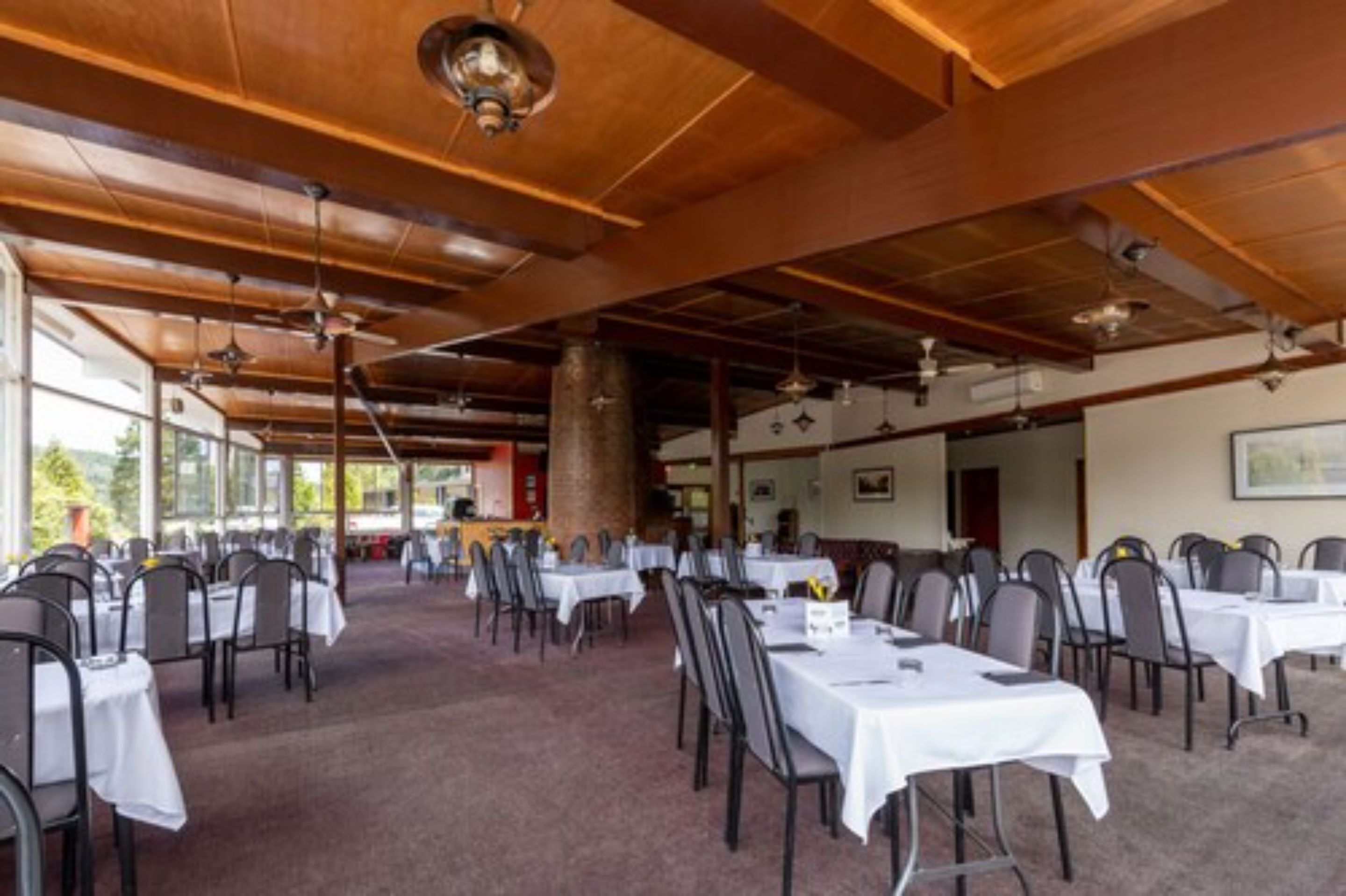 A spacious dining area with multiple tables and chairs, wooden ceiling beams, and large windows allowing natural light.
