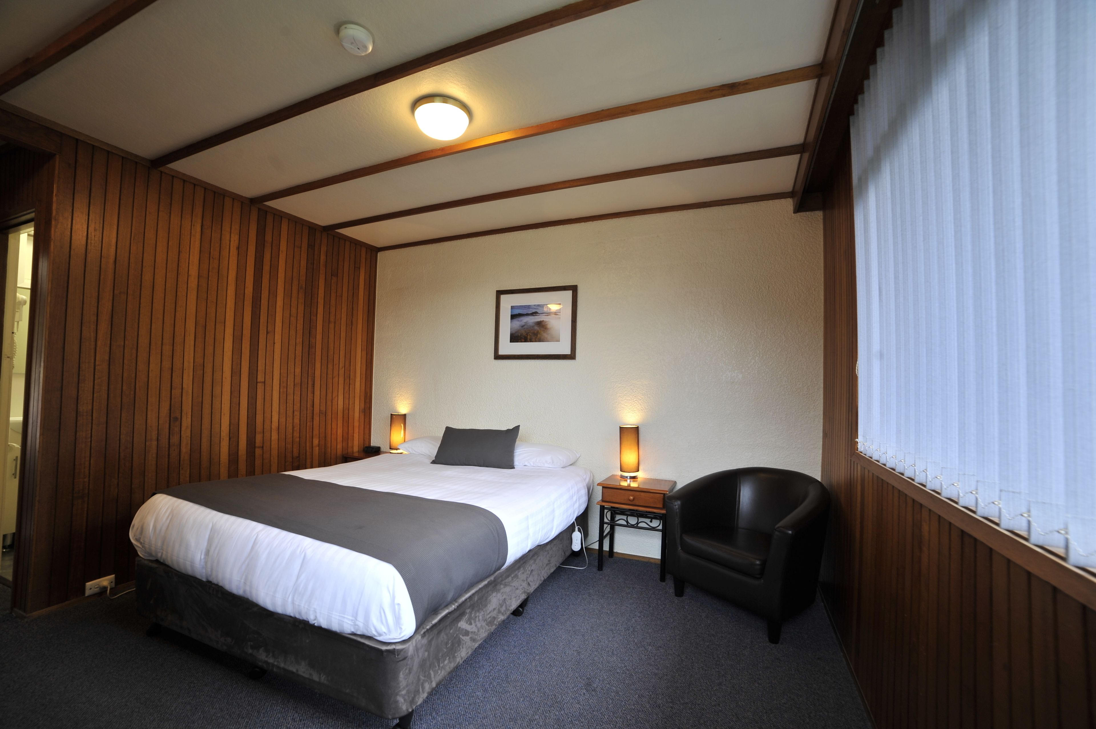 A hotel room with a large bed, bedside lamps, a small table, a chair, and a picture on the wall.