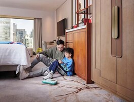 A hotel room with a bed, a city view, a wooden wardrobe, and a television.
