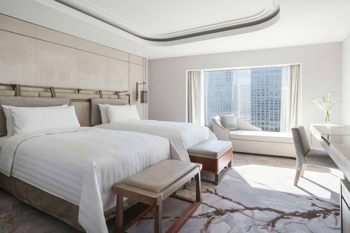 A modern hotel room with a large bed, a seating area with a bench, a desk, and a view of the cityscape through a large window.