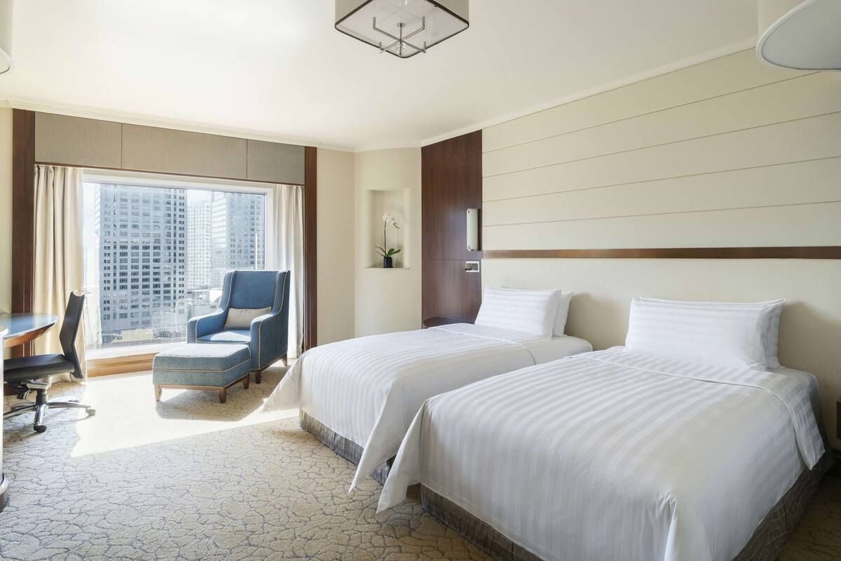 A hotel room with two beds, a desk, a chair, a large window with a view of buildings, and a ceiling light fixture.